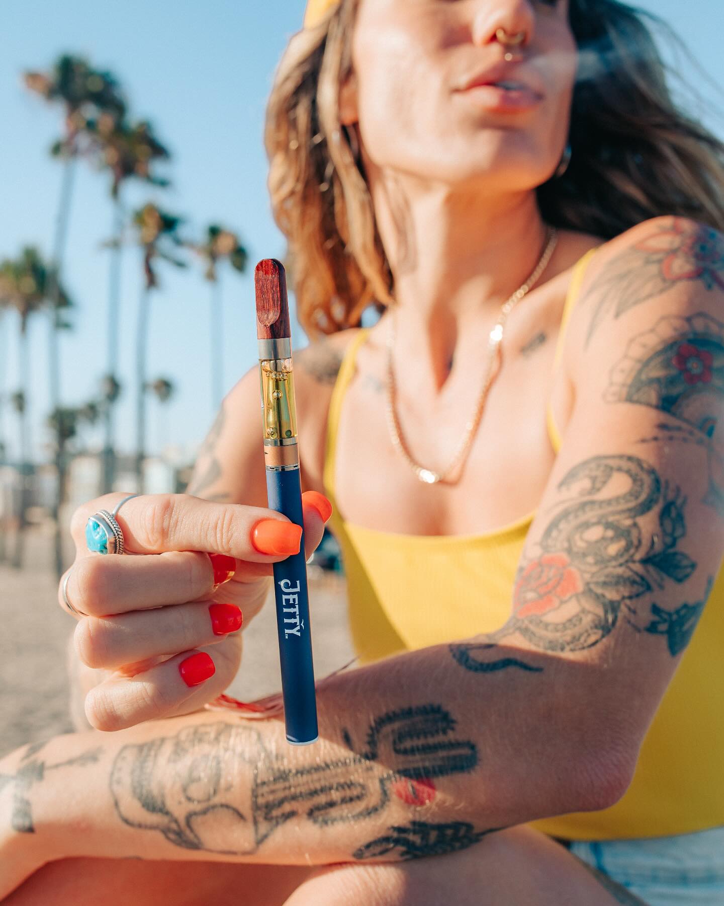 Tattooed woman in a yellow top holding a Jetty vape pen and cartridge on a palm lined beach