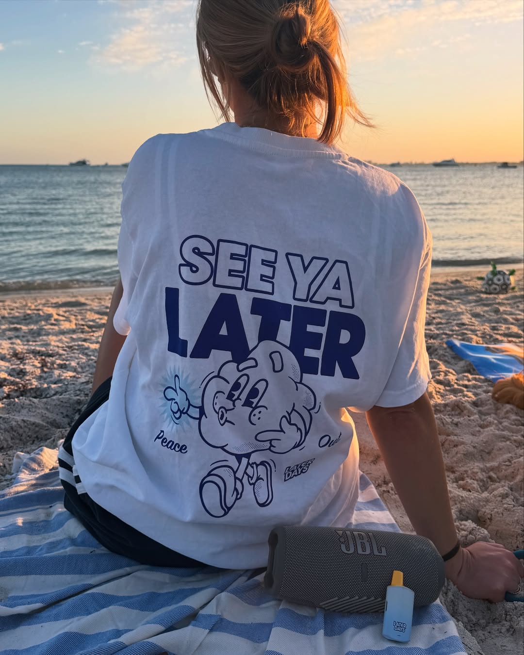 Woman in a See Ya Later tee sitting on a striped beach towel at sunset beside a Later Days vape