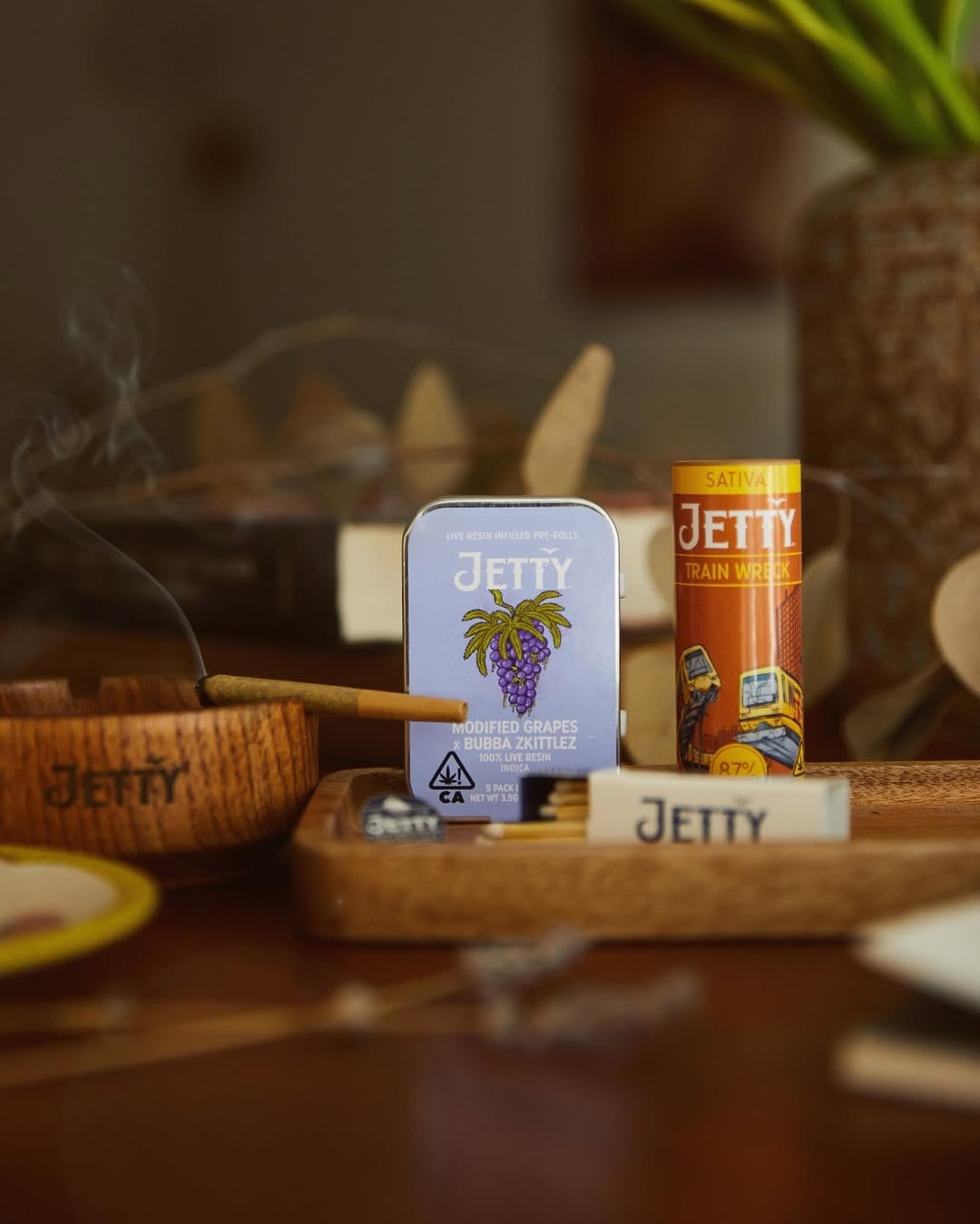 Lit Jetty pre-roll on a wooden ashtray beside a Modified Grapes tin and Train Wreck cartridge tube
